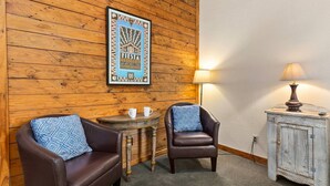 Una habitación con paredes de paneles de madera, dos sillones de cuero con cojines estampados, una mesita de madera con una lámpara y un cartel enmarcado en la pared.