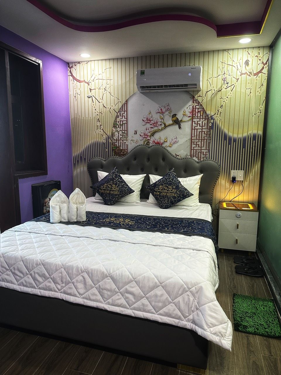 Un dormitorio con cama, almohadas, mesita de noche, aire acondicionado y una pared con estampado floral.