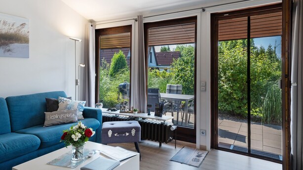 Ein modernes Wohnzimmer mit einem blauen Sofa, einem Couchtisch mit Blumen in einer Vase, einem Heizkörper und einer Schiebetür zu einem Außenbereich mit Tisch und Stühlen.