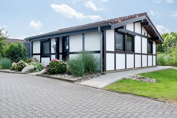 A modern two-story house with a brick roof, white walls, and dark trim. It features a paved driveway and a well-maintained garden with various plants and flowers.