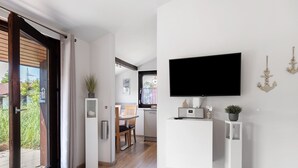Ein modernes Interieur mit einem an der weißen Wand befestigten Flachbildfernseher, einem Essbereich mit Holztisch und -stühlen sowie einer Küche im Hintergrund.