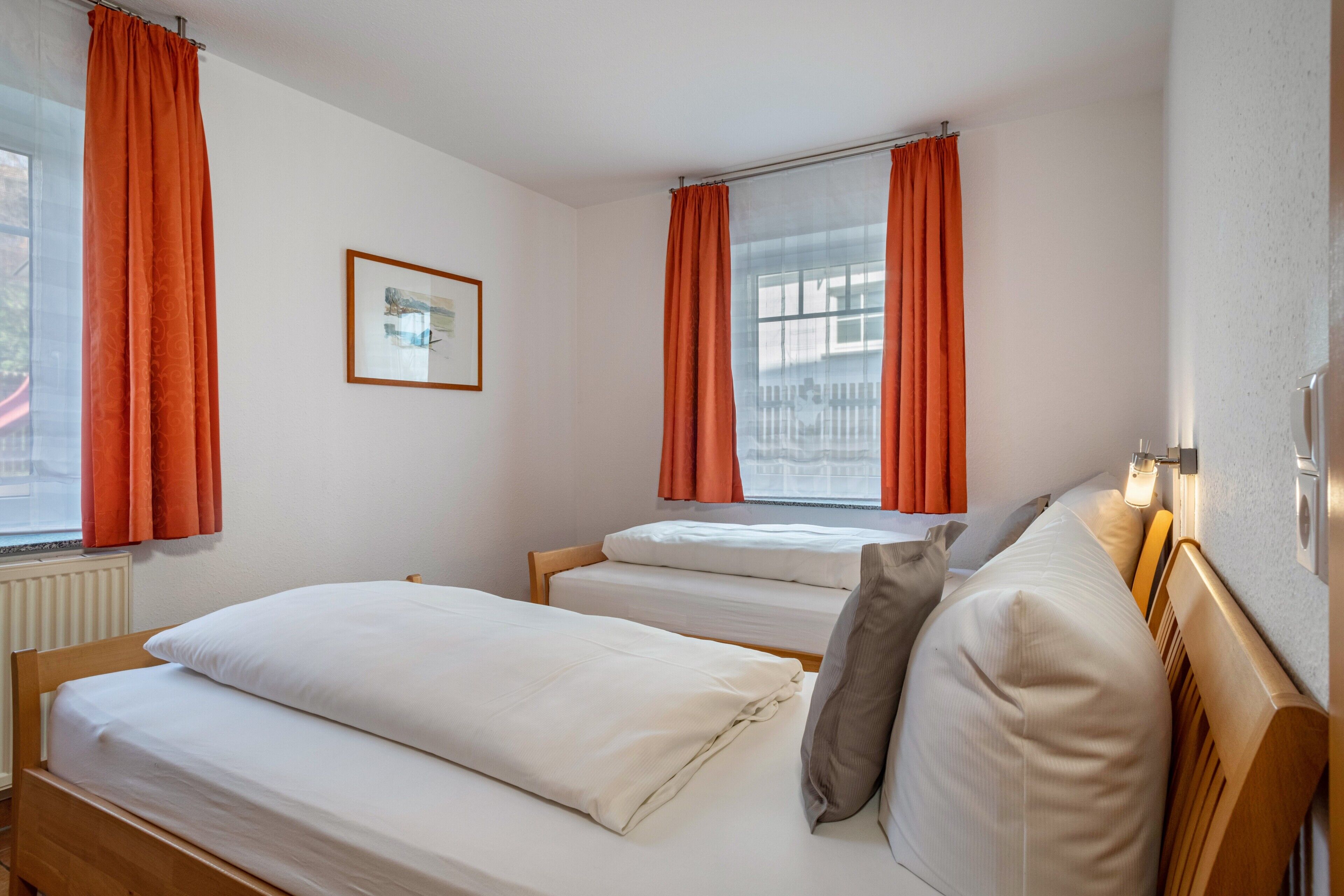 Une chambre avec deux lits, des rideaux orange, un tableau encadré au mur et une fenêtre donnant sur des immeubles.
