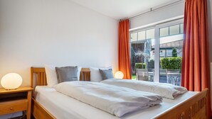 Uma cama de casal com roupa de cama branca, uma cabeceira de madeira, uma mesa de cabeceira com abajur e uma janela com cortinas laranja.
