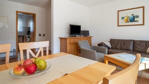 Uma sala de estar com sofá, televisão em um móvel, mesa de jantar com maçãs e um quadro na parede.