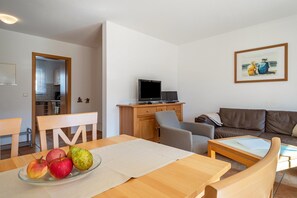 Una sala de estar con un sofá, un televisor sobre un mueble, una mesa de comedor con manzanas y un cuadro en la pared.