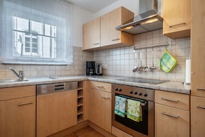Una cocina con armarios de madera, lavavajillas de acero inoxidable, horno empotrado y ventana con cortinas translúcidas.