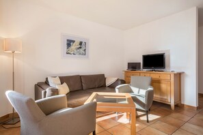 Un salon moderne comprenant un canapé, deux fauteuils, une table basse, un téléviseur posé sur un meuble en bois et un lampadaire.
