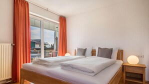 Een tweepersoonsbed met witte lakens, een houten hoofdbord, een nachtkastje met een lamp en uitzicht naar buiten door een groot raam met oranje gordijnen.