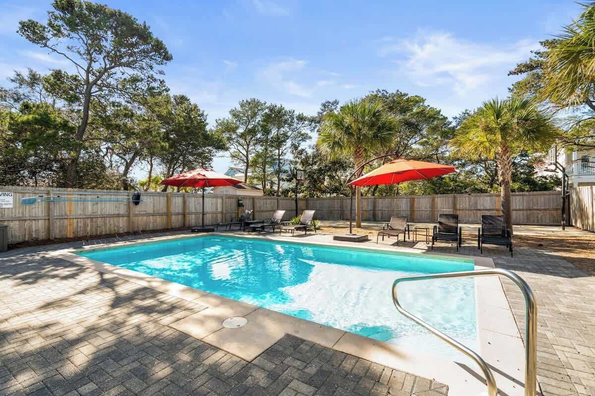 A swimming pool with lounge chairs and umbrellas, surrounded by trees and a wooden fence.