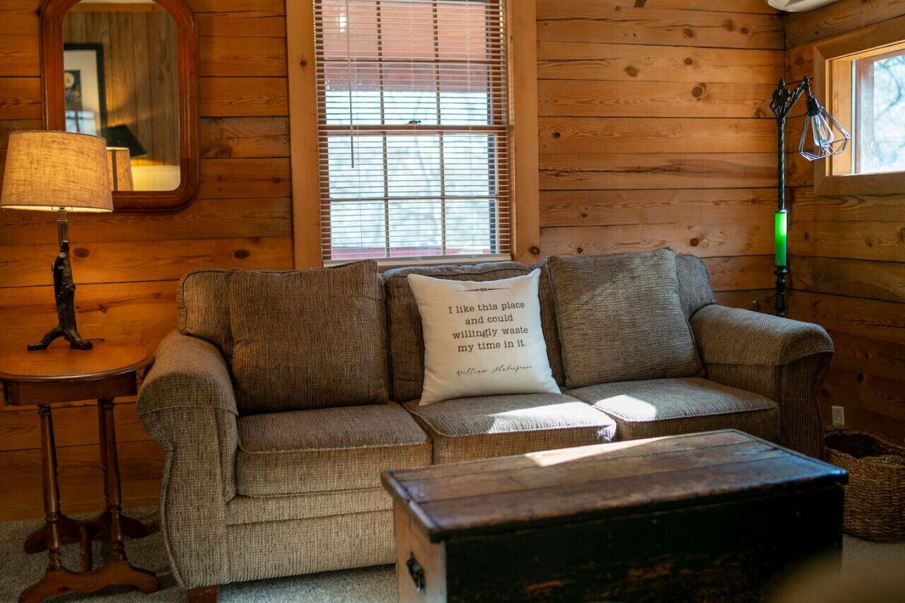Ein gemütliches Zimmer mit Holzwand, einer Couch mit Kissen, einer Lampe und einem Fenster mit Jalousien.