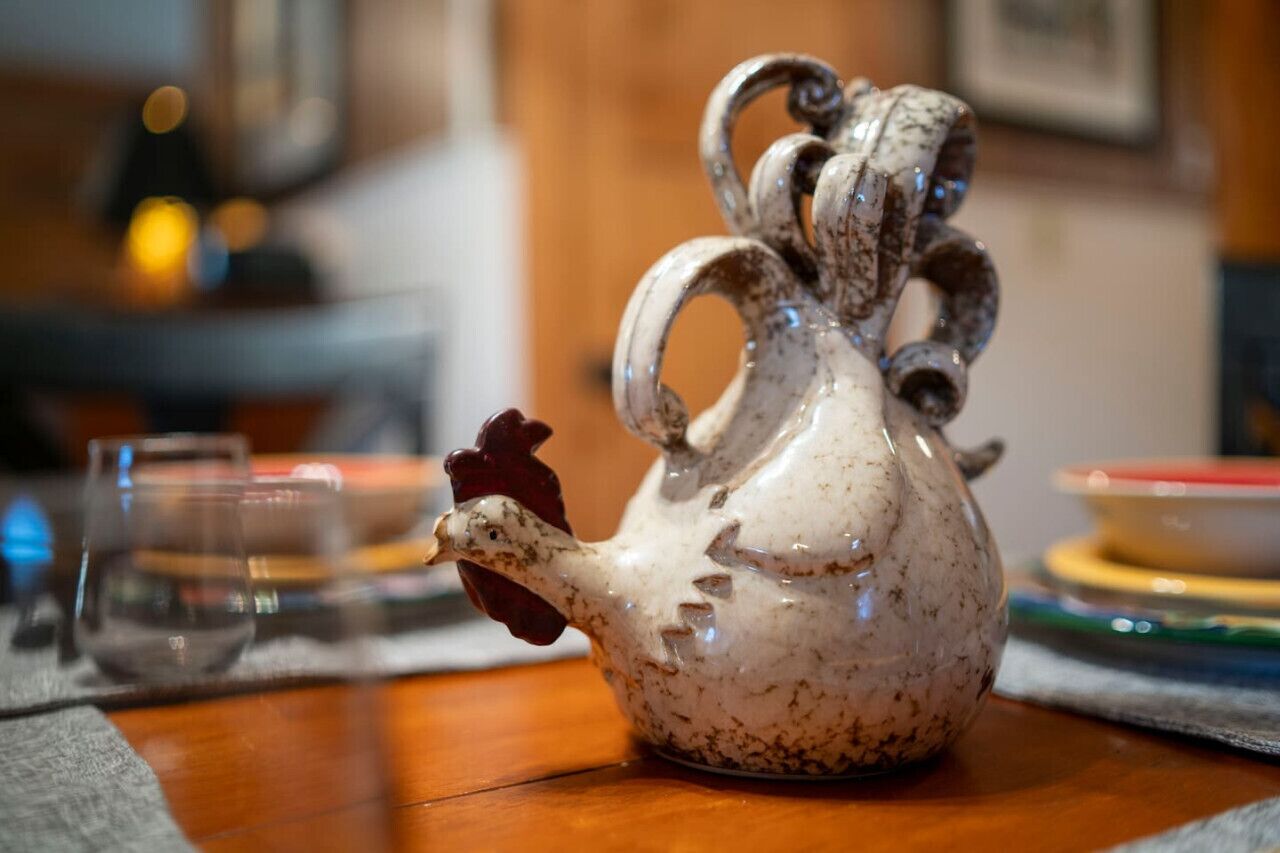 Un pichet en céramique en forme de coq est posé sur une table en bois, avec un verre et une bougie en arrière-plan.