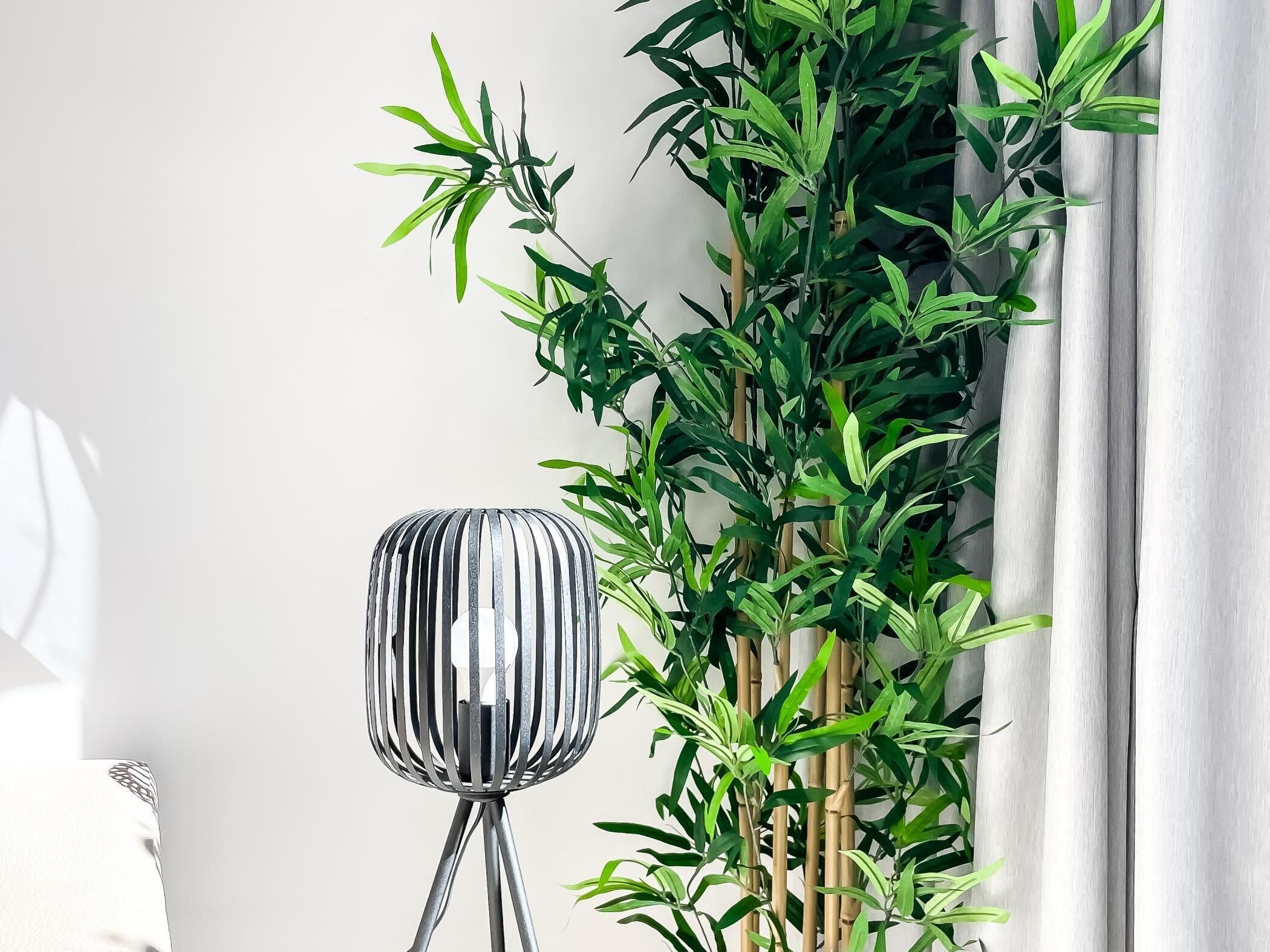 A modern lamp with a cylindrical base and vertical slats, positioned next to a bamboo plant with green leaves against a white wall.