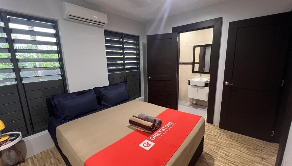 A hotel room with a bed, a nightstand, a window with blinds, an air conditioning unit, and a door leading to a bathroom.