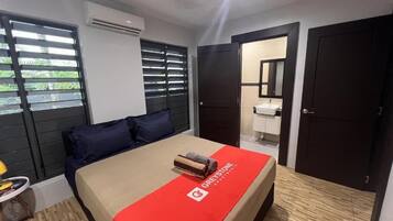A hotel room with a bed, a nightstand, a window with blinds, an air conditioning unit, and a door leading to a bathroom.