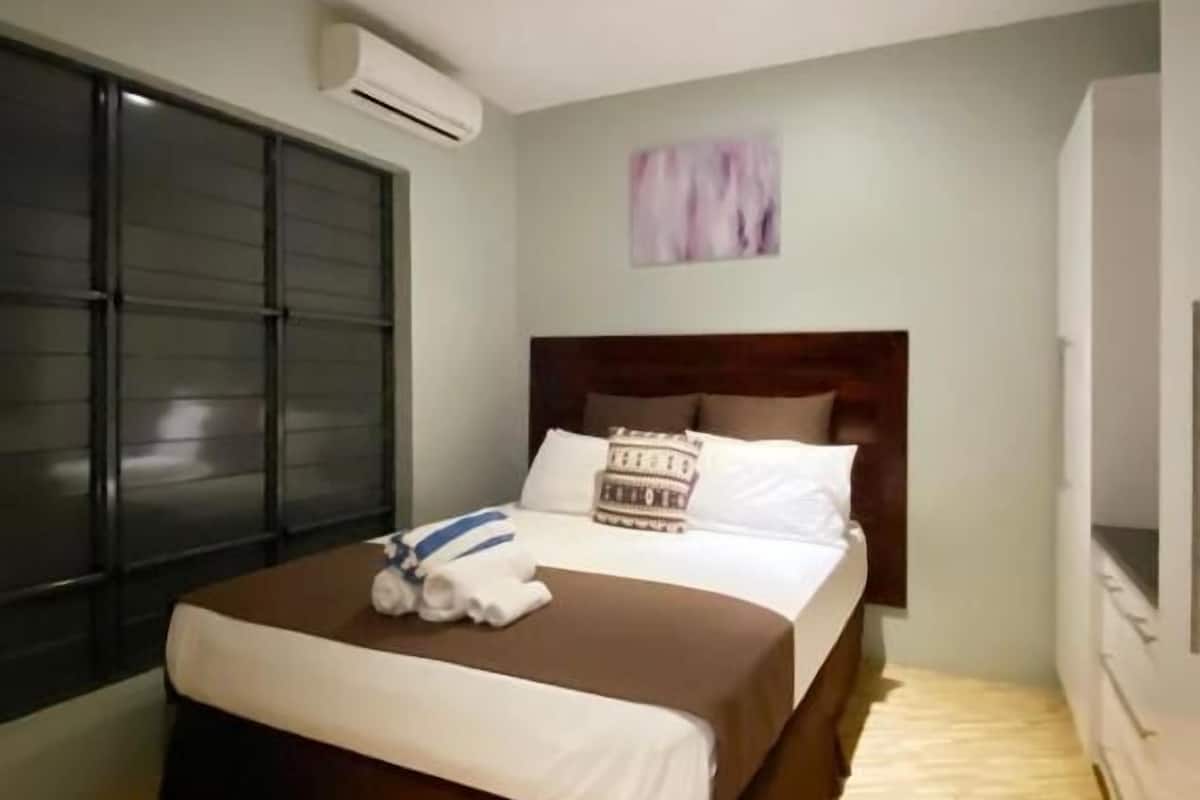 A bedroom with a bed, a ceiling fan, an air conditioning unit, a window, and a painting on the wall.