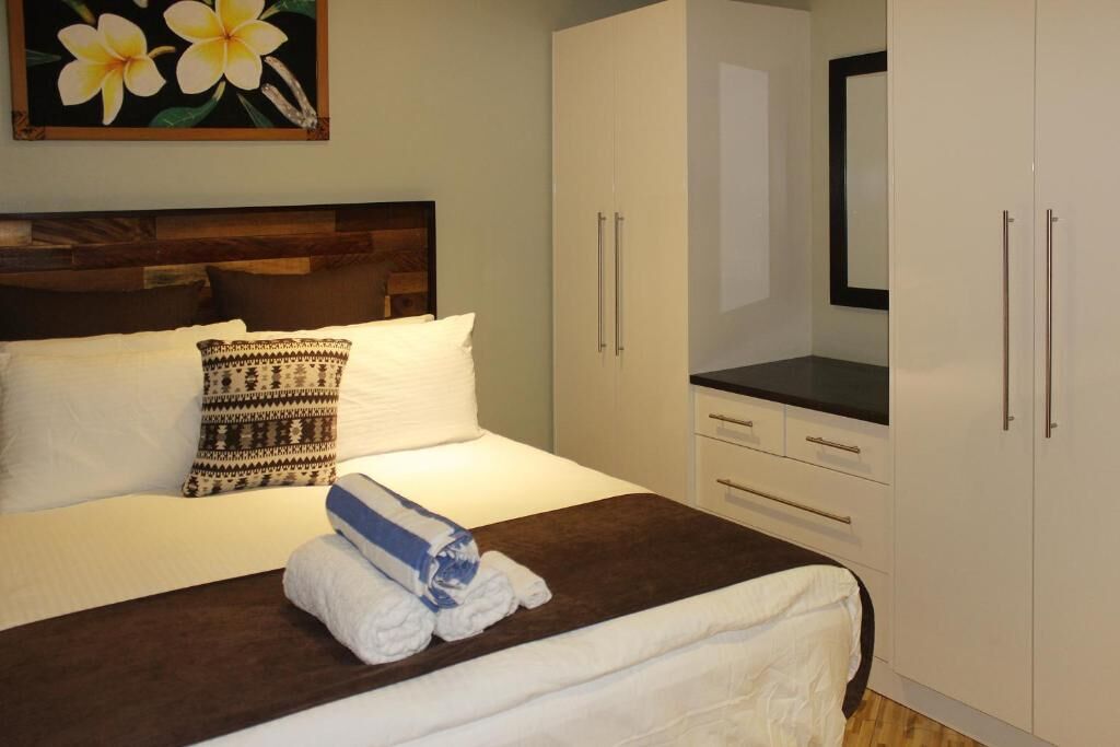 Letto rifatto con lenzuola bianche, cuscino decorato e asciugamani piegati. Si nota una testiera in legno, un'armadio bianco con maniglie argentate e un piano d'appoggio nero.
