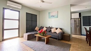 A living room with a sofa, a coffee table with flowers, a grey rug, wooden flooring, a ceiling fan, an air conditioning unit, a kitchen area with a refrigerator, and a window with blinds.