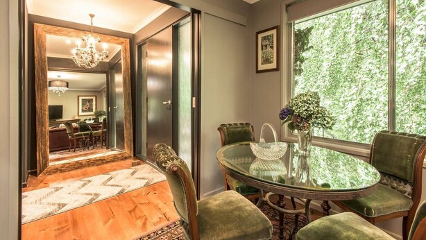 Un espace repas avec une table en verre et des chaises recouvertes d’un tissu vert, un lustre et une vue sur la verdure grâce à de grandes fenêtres.