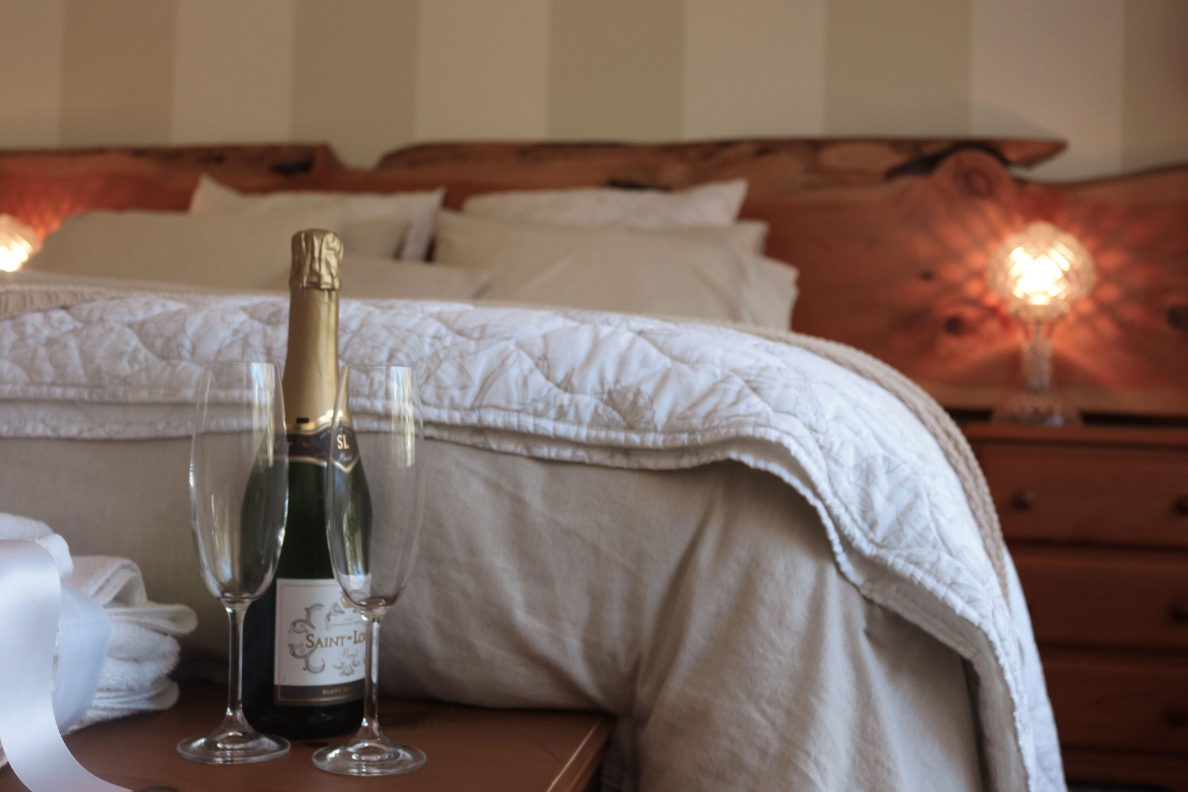 A bed with a wooden headboard, a bottle of champagne, and two glasses on a bedside table.