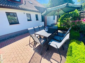 En udendørs patio med et bord og stole, en belagt terasse med mursten og en hvid bygning med vinduer.