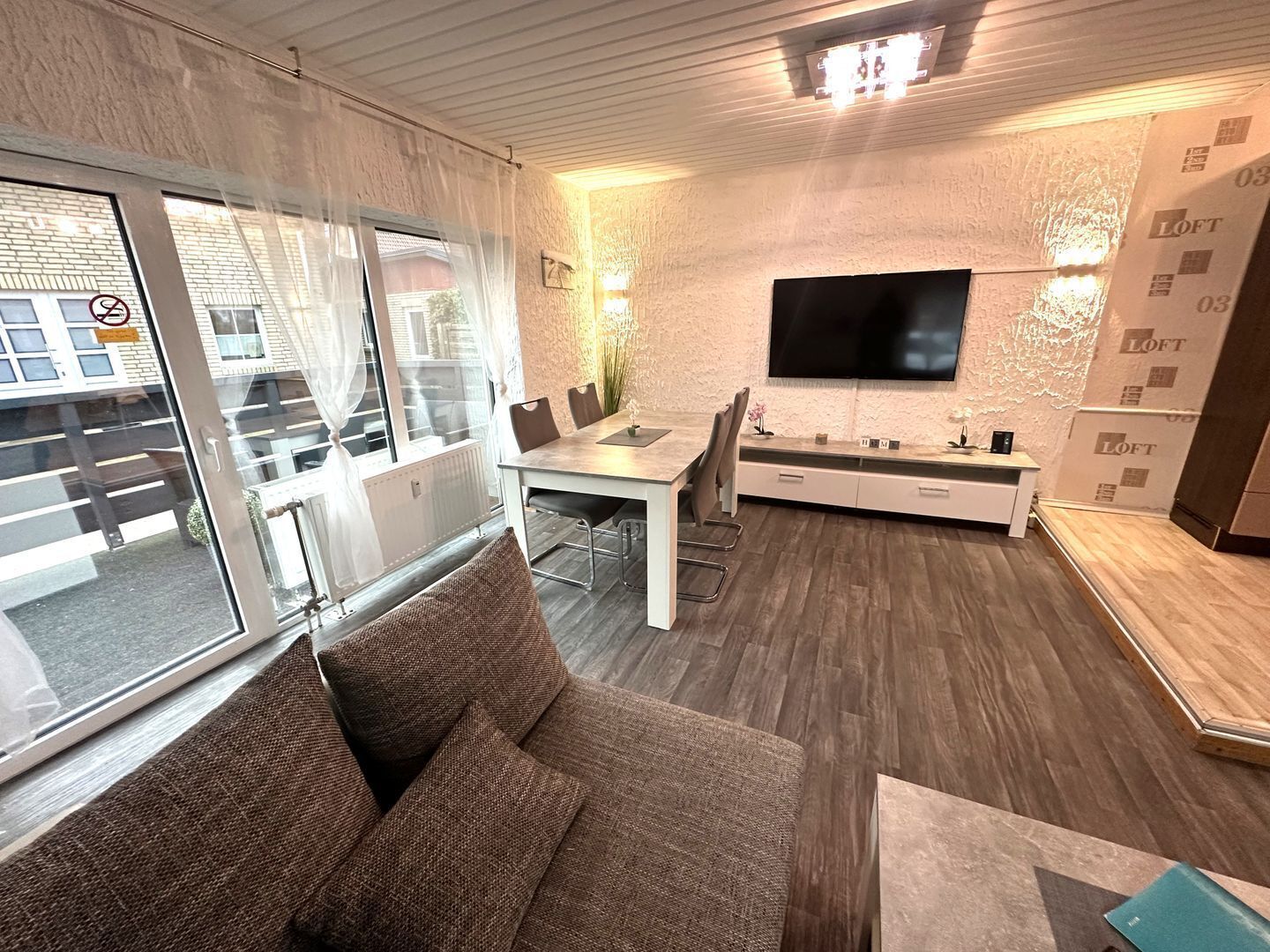 A modern living room with a sofa, a dining area with a table and chairs, a flat-screen TV mounted on the wall, and a large window with curtains.