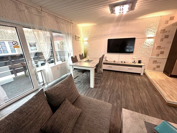 A modern living room with a sofa, a dining area with a table and chairs, a flat-screen TV mounted on the wall, and a large window with curtains.