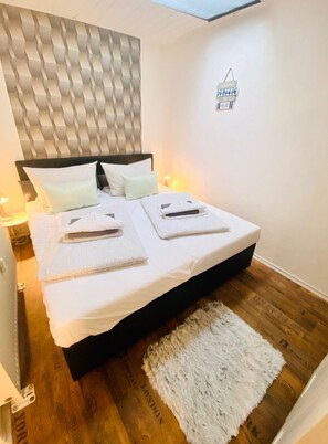 A modern bedroom with a wooden floor, a fluffy rug, a bed with white linens and towels, a headboard with a textured pattern, and a wall-mounted sign.