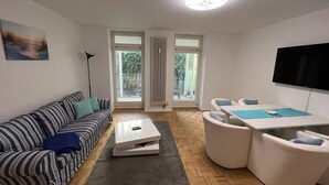 A modern living room with a striped sofa, a white coffee table, and a dining area with white chairs and a table. - Apartment Bernstein - Ferienwohnung Bernstein Ruhig mit Garten (Rostock)
