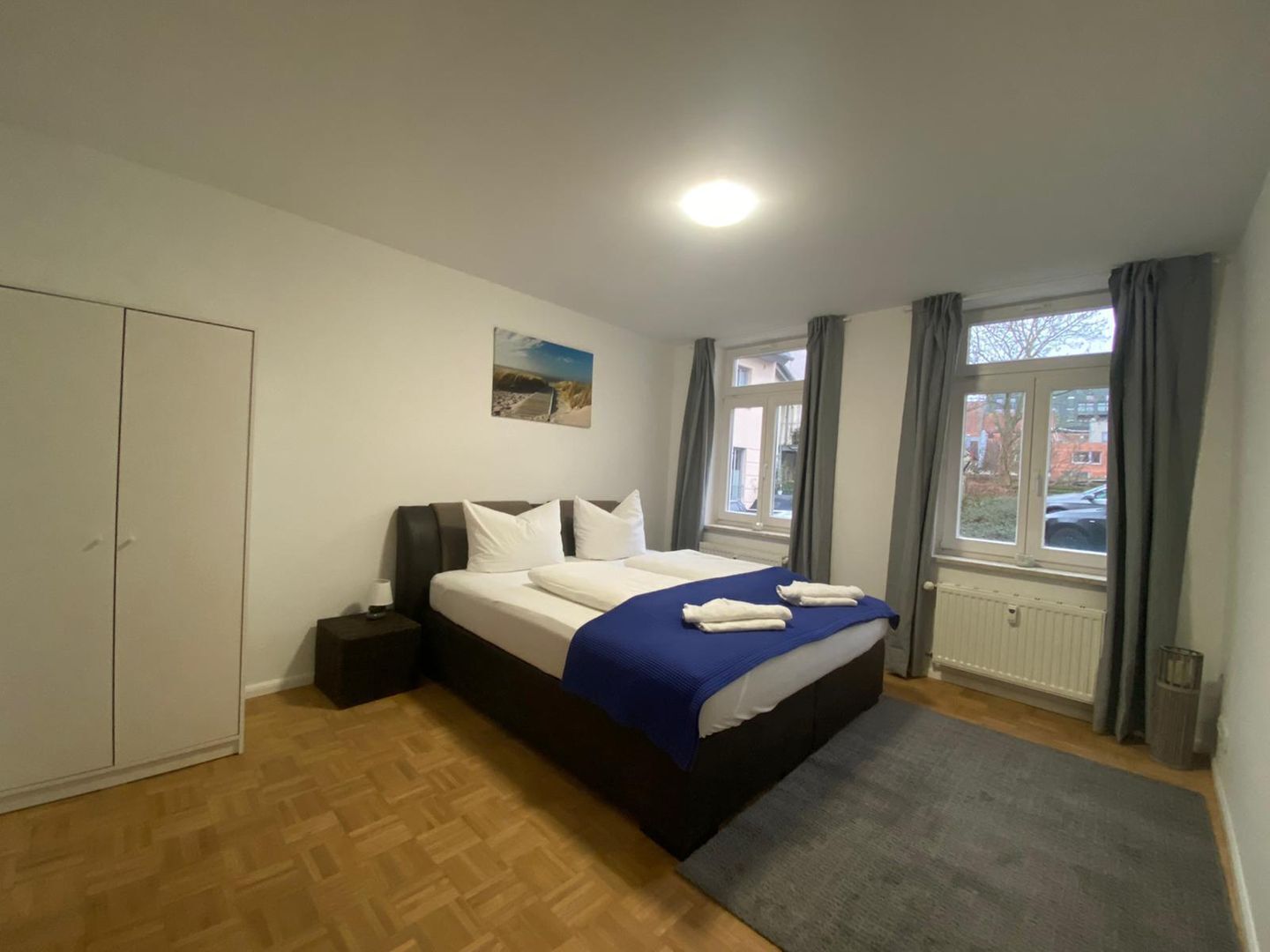 Un dormitorio con cama, mesita de noche, ventana con cortinas, radiador y puerta.