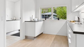 A modern kitchen with white cabinetry, a window with a view of a house, and a countertop with a kettle and a knife block. - "Majvor" - 200m from the sea by Interhome (Hadsund)