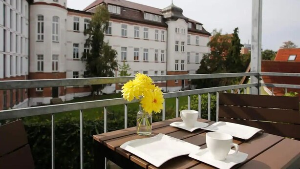 A balcony with a table set for two, a vase with yellow flowers, and a view of a large building with multiple windows and a dome.