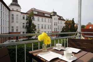 A balcony with a table set for two, a vase with yellow flowers, and a view of a large building with multiple windows and a dome.
