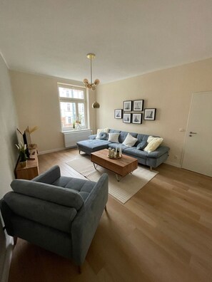 A spacious living room with a blue sofa, a coffee table, and a grey armchair. There is a window with a view of buildings outside.