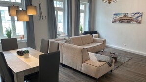 Ein modernes Wohnzimmer mit einem beigefarbenen Sofa, einem Esstisch und Stühlen. Es gibt eine Chandelieber, ein Gemälde vom Eiffelturm und eine Topfpflanze.