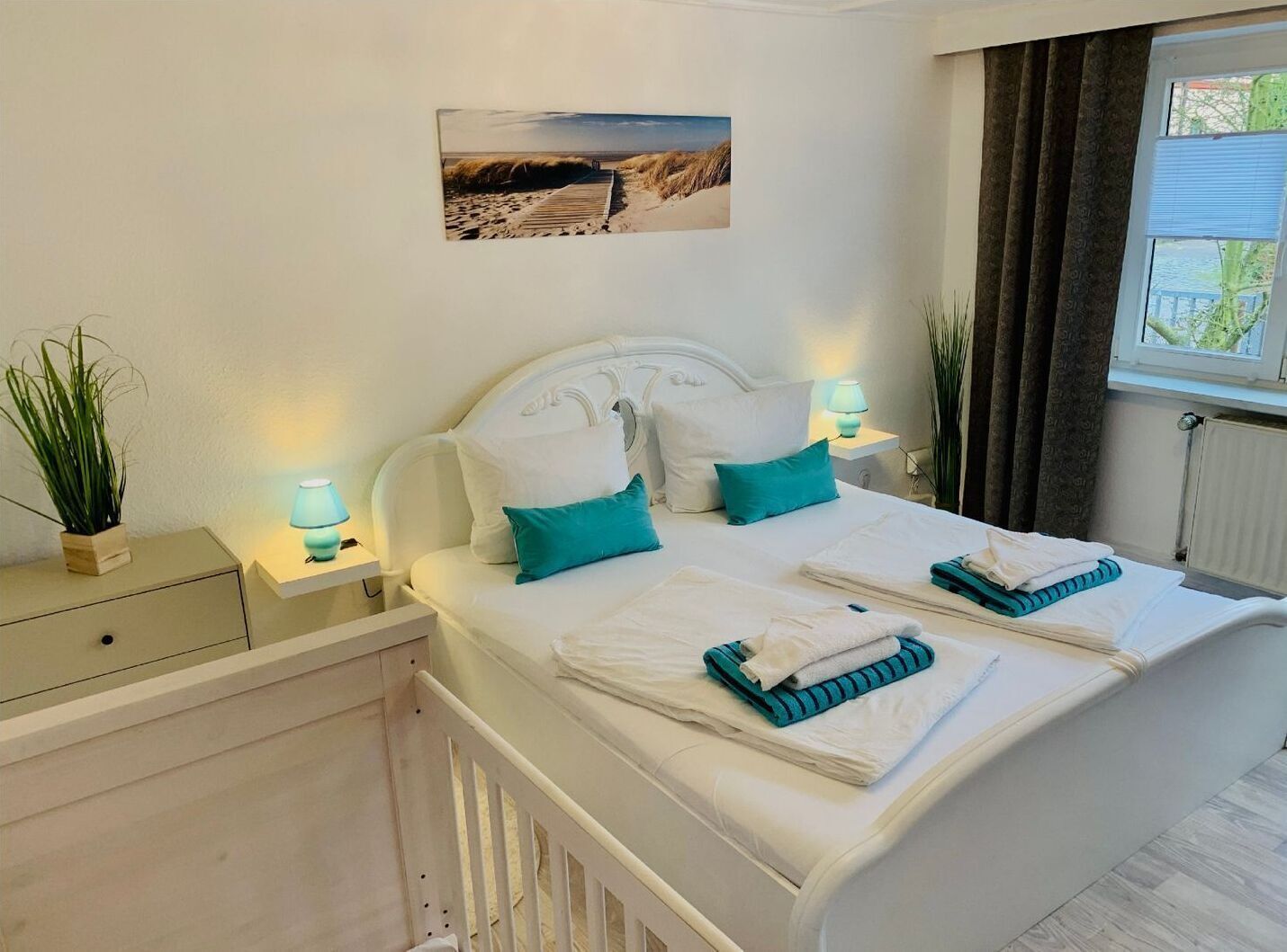 Een net opgemaakt bed met kussens en opgevouwen handdoeken, een nachtkastje met een lamp, een potplant en een ingelijst foto aan de muur.