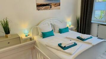 Een net opgemaakt bed met kussens en opgevouwen handdoeken, een nachtkastje met een lamp, een potplant en een ingelijst foto aan de muur.