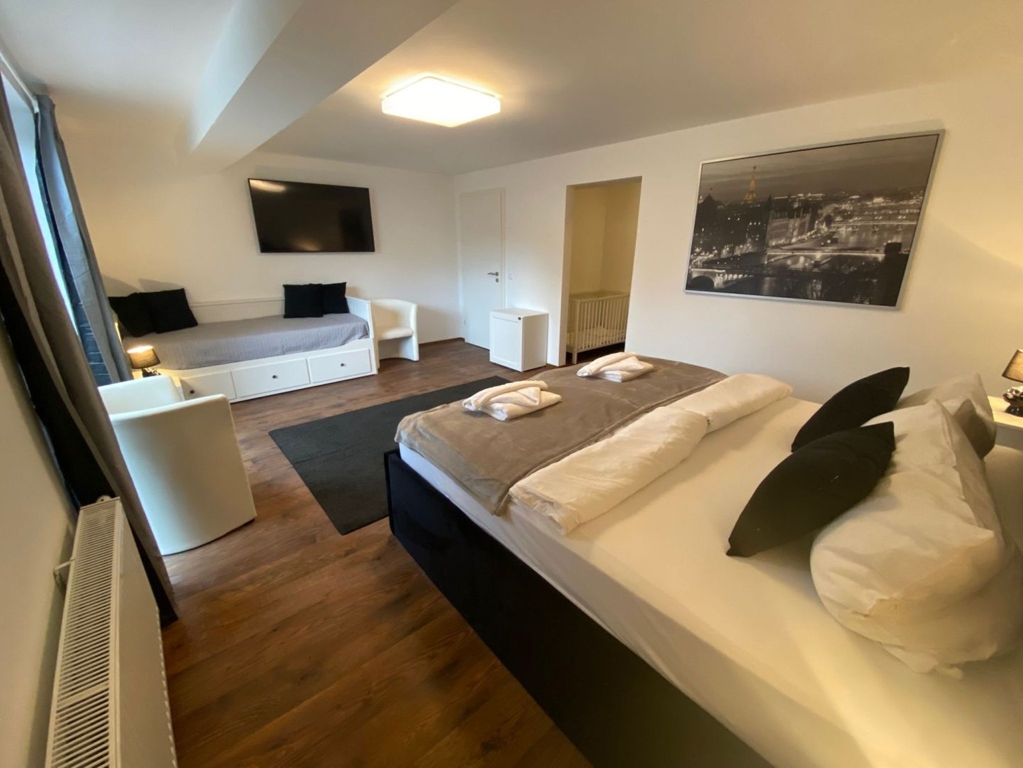 Ett modernt sovrum med en stor säng, ett skrivbord, en stol, en tv och en tavla som föreställer en stadsbild.