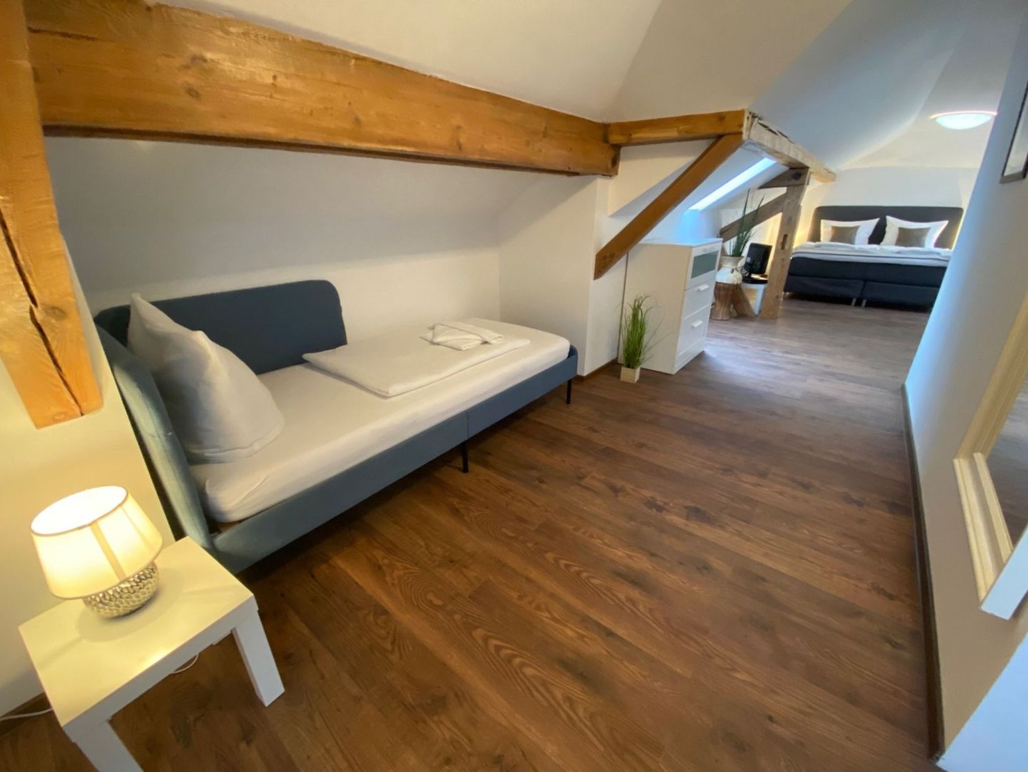 Dormitorio estilo loft con cama, mesita de noche, lámpara, vigas de madera y vista a otra habitación.