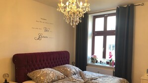 Un dormitorio con cabecera acolchada, ropa de cama estampada, lámpara de noche, ventana con cortinas y un adorno de pared con texto.