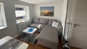 Ett kompakt vardagsrum med en soffa, ett litet bord, ett fönster med persienner, en tavla på väggen och en dörr.