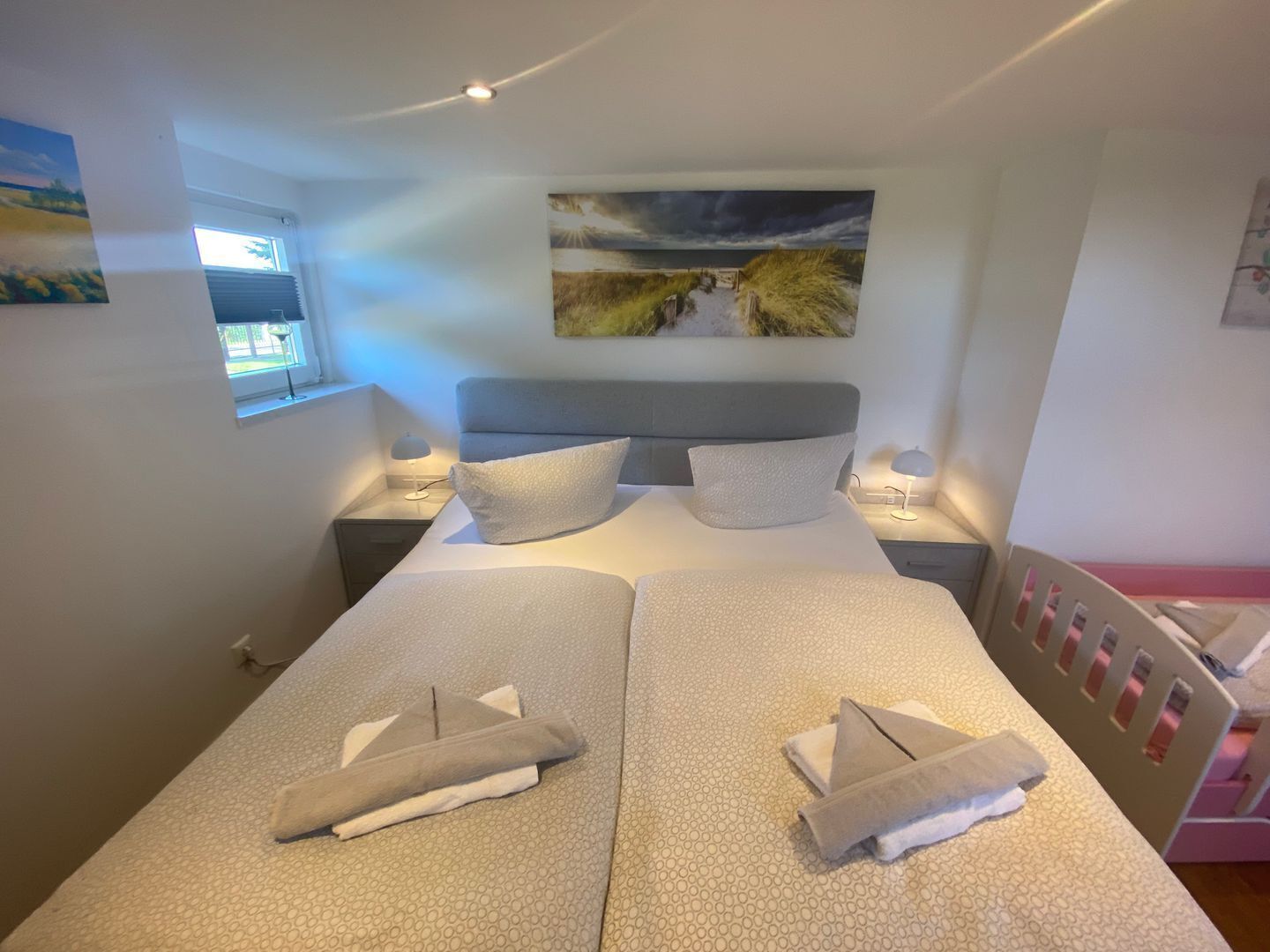 Ein Doppelbett mit zwei Kissen, ein Nachttisch mit Lampe, ein Bild an der Wand und ein Fenster mit Jalousien.