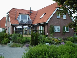 Une maison à deux étages, avec un toit recouvert de tuiles rouges, un balcon et un jardin où poussent différentes plantes et fleurs.