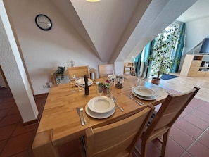 Un coin repas avec une table en bois dressée pour quatre personnes, une horloge au mur et une vue sur l’extérieur.