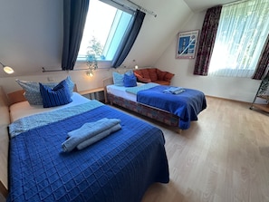 Une chambre avec deux lits, chacun agrémenté de draps et d’oreillers bleus, une petite table avec une lampe et une fenêtre avec des rideaux.