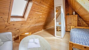A wooden room with a sloped ceiling, a bed, a chair, a small table, and a window.