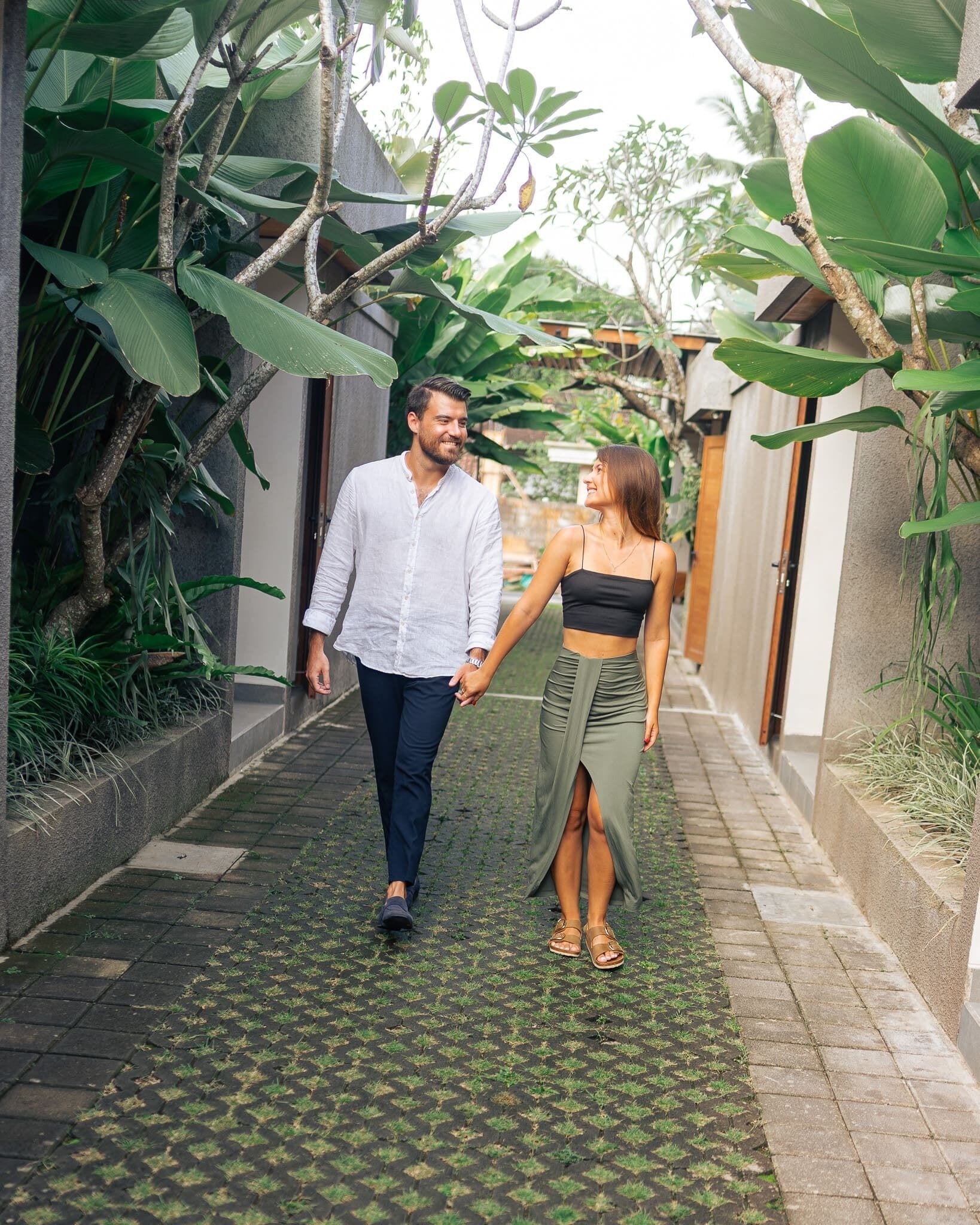 A couple walking hand in hand down a narrow, paved pathway flanked by lush greenery and stone walls.