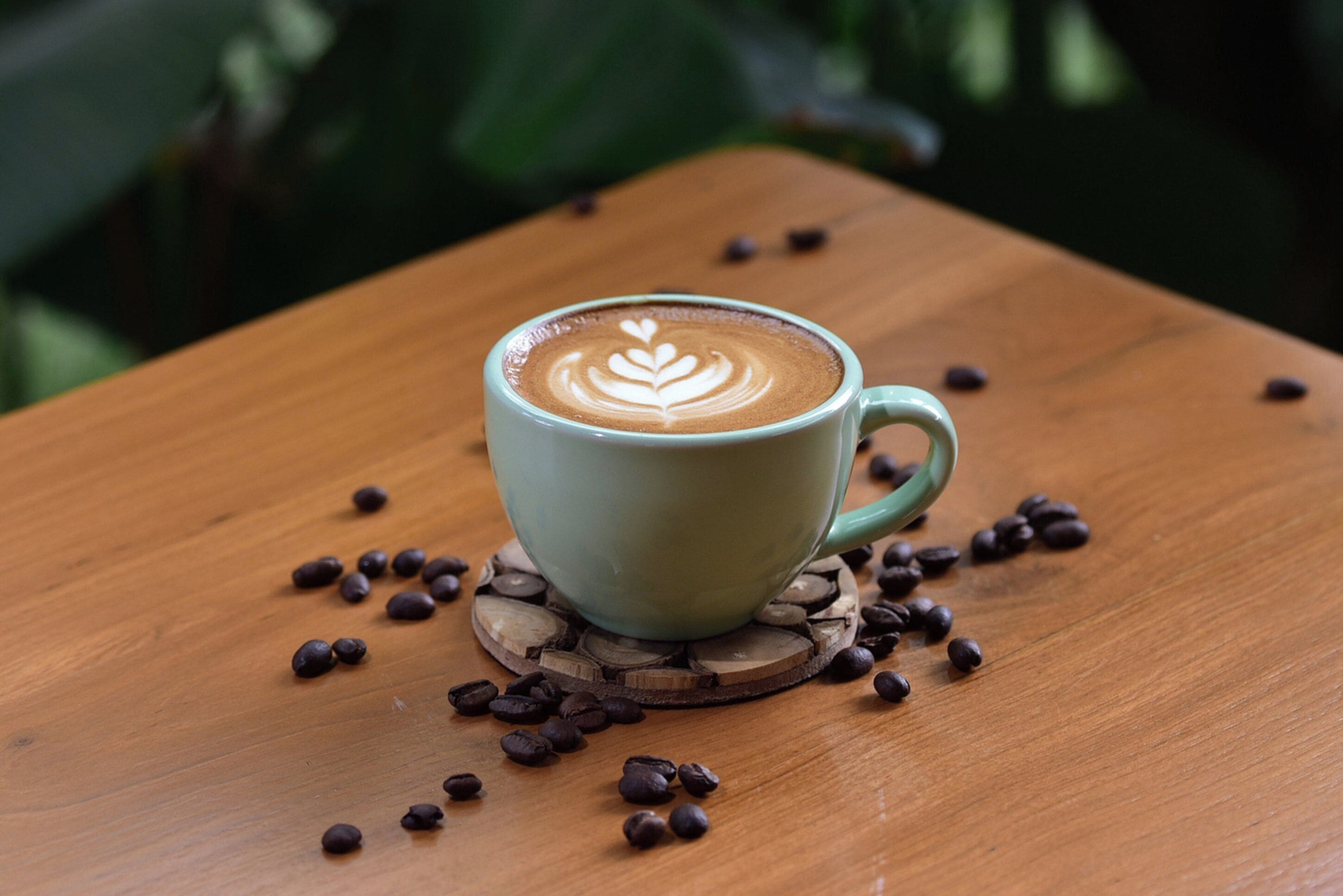 A cup of coffee with latte art on a wooden coaster, surrounded by scattered coffee beans on a wooden table.
