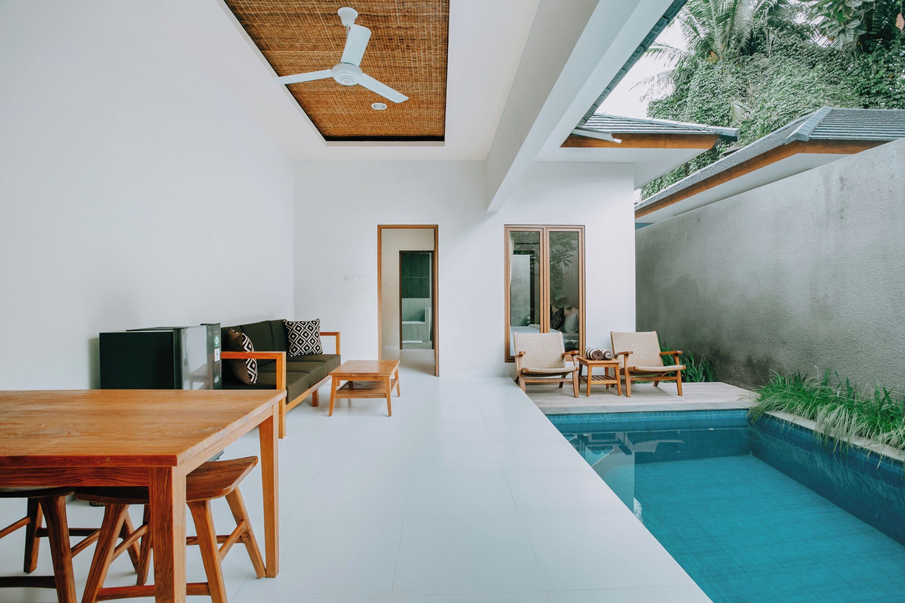 Un salon moderne avec une table en bois, des chaises, un canapé et un téléviseur. Une porte vitrée donne accès à une autre pièce, et il y a un espace piscine avec des chaises longues.