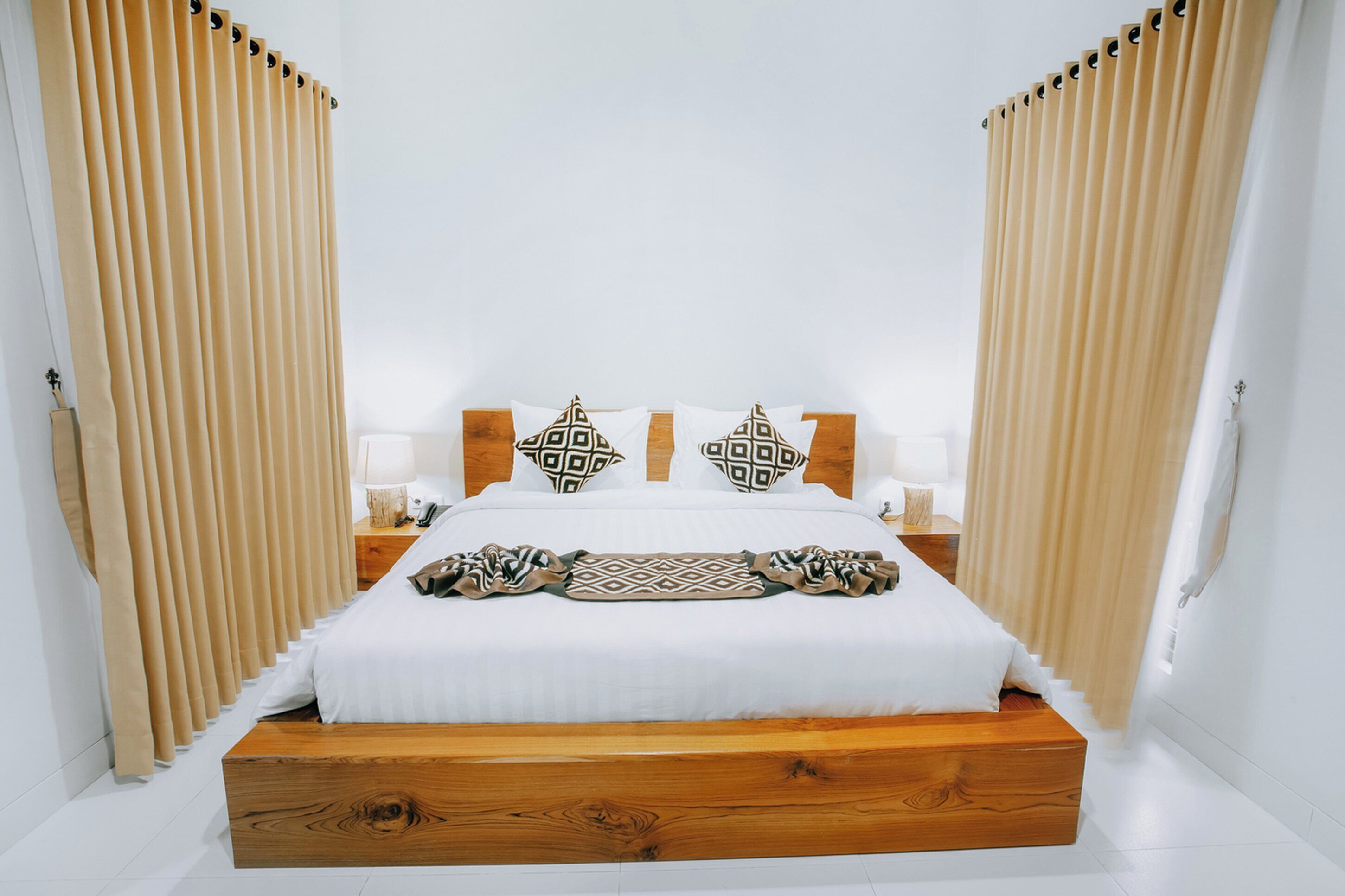 A bedroom with a wooden bed, white bedding, patterned pillows, and beige curtains.