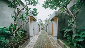 A narrow pathway flanked by concrete walls and lush greenery, leading to a building with wooden doors. - Mahayoga Ubud Private Pool Villa Spa And Resto (Ubud)